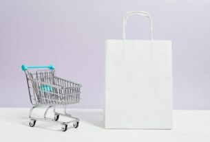 Minimalist image of a white shopping bag and mini cart on a soft pastel background.